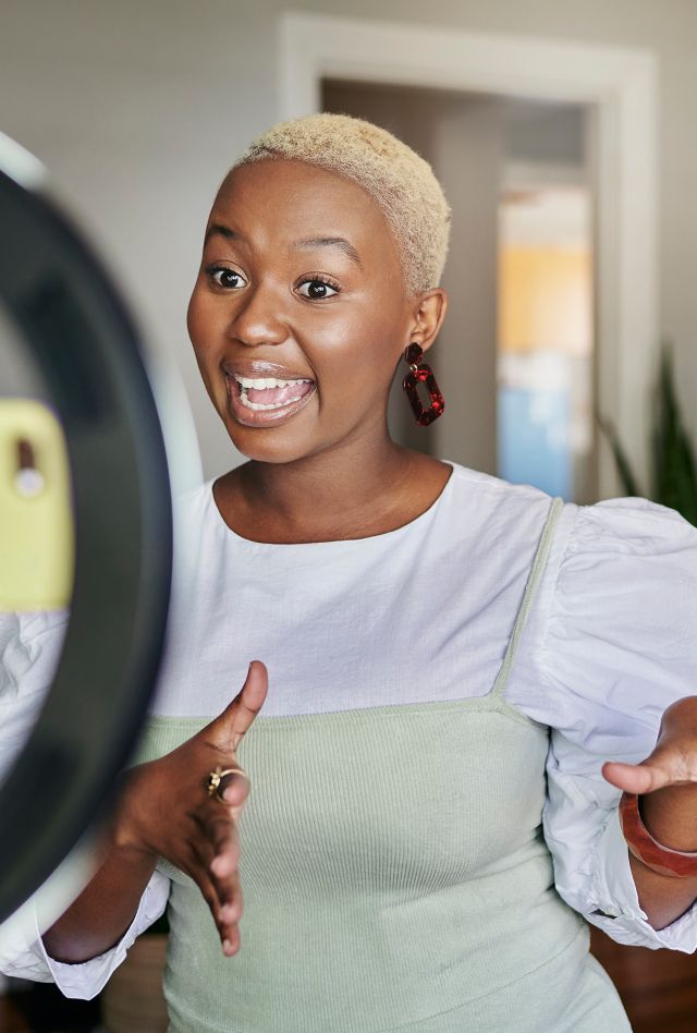 Smiling young dark-skinned female animatedly speaking to a smart phone connected to a ring light.