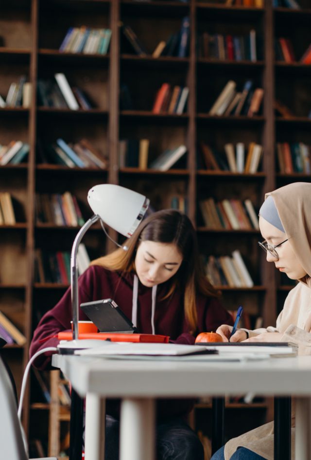 Serious young diverse female students writing while sitting at desk in light modern library