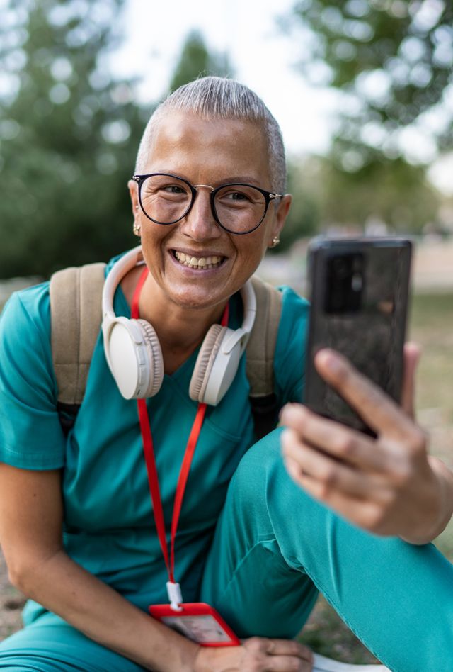 Smiling female healthcare worker in scrubs taking a selfie or having a video call on her smartphone while relaxing outdoors in a park