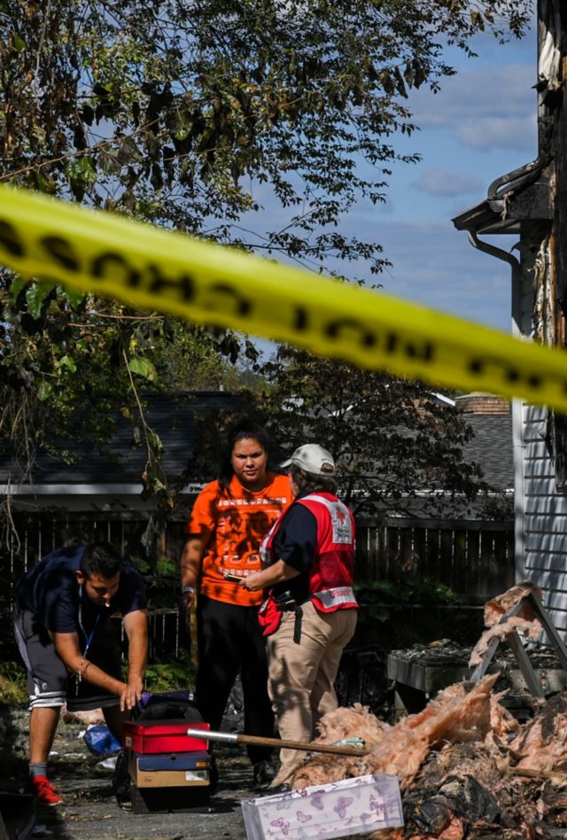 A woman wearing a Red Cross vest speaks to someone outside of a house with serious fire damage
