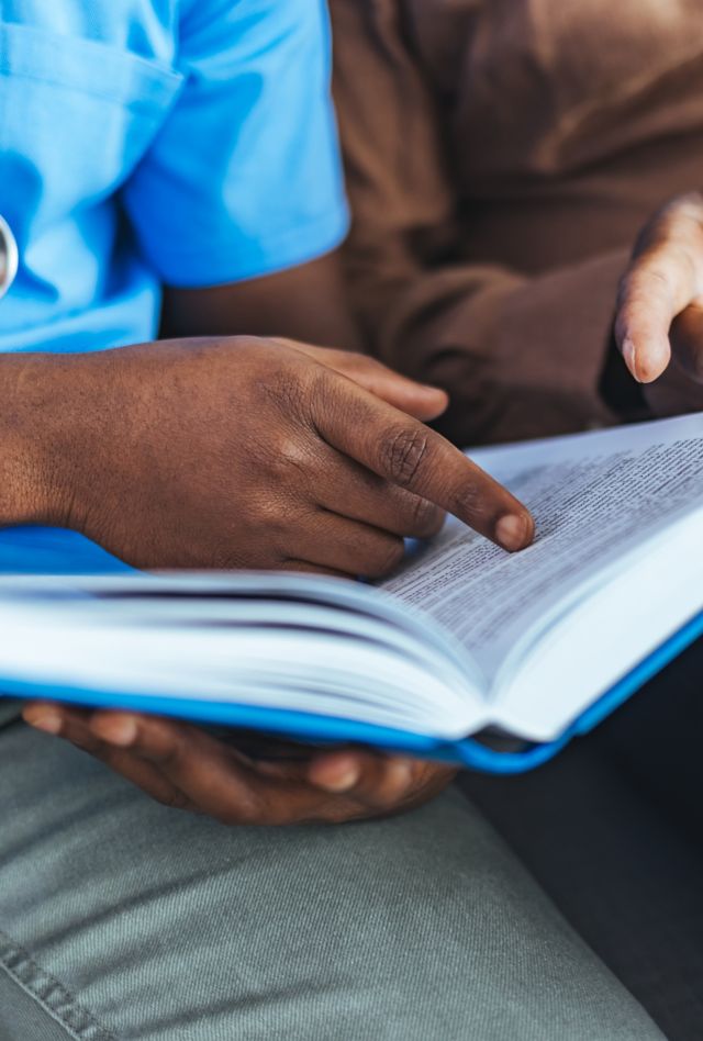 Senior elderly man reading book with nurse while sitting on sofa.