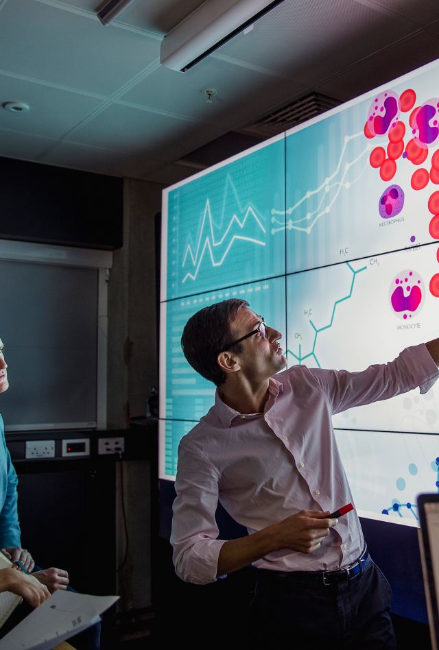 Group of business professionals in a dark meeting room, standing in front of a large screen with scientific displays of DNA and cells being presented.