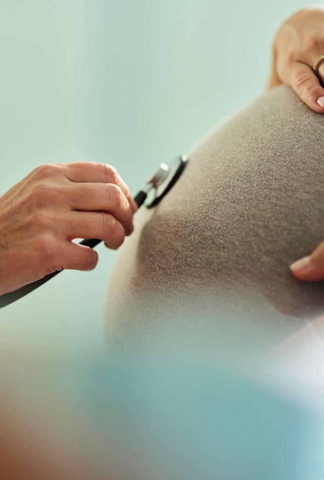 Close up of doctor using stethoscope on a person's pregnant stomach. Doctor has white lab coat, person have gray shirt