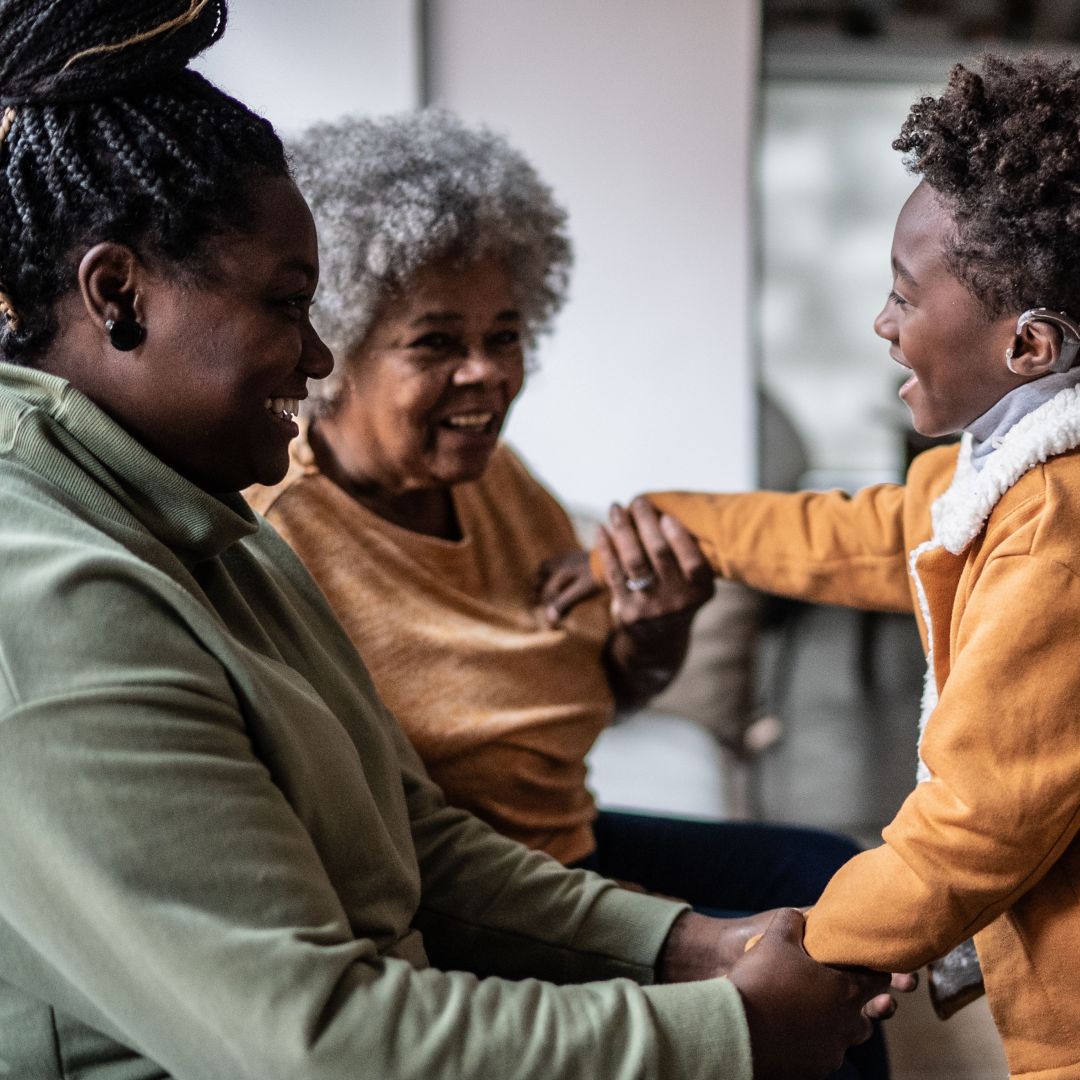 A child wearing a hearing aid plays with his mother and grandmother at home