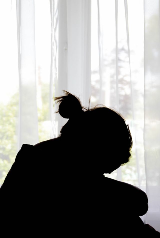 Silhouette of a person in front of a window. Their arms are wrapped around their knees and their head is bent.