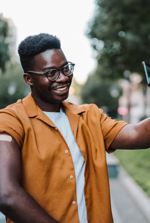Shot of young smiling man taking selfie after receiving vaccine. black male in an orange short sleeve button down shows his shoulder with bandaid.