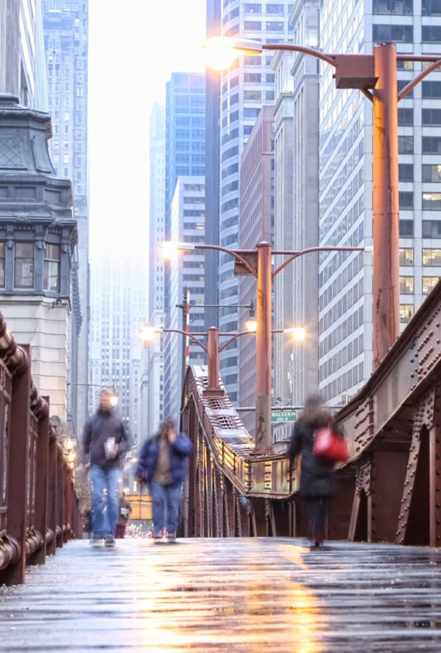 Pedestrians crossing the LaSalle Street Bridge in Chicago on a rainy day, with skyscrapers visible in the background.