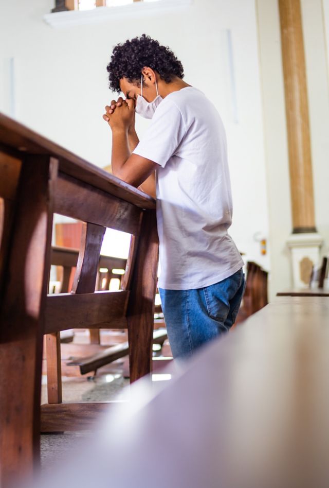 Young man, wearing a white protective mask, inside the church, praying in times of coronavirus