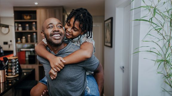 Smiling, dark-skinned man carrying young boy with braids on his back.