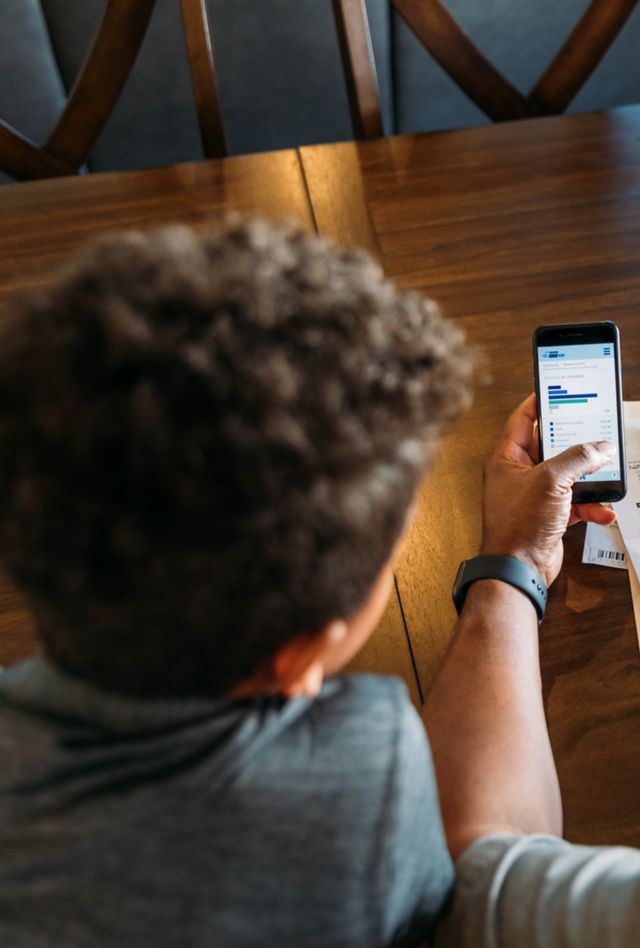 A middle-aged man sits at his dining room table with his young son teaching him about home finances. He is working from home and telecommuting.