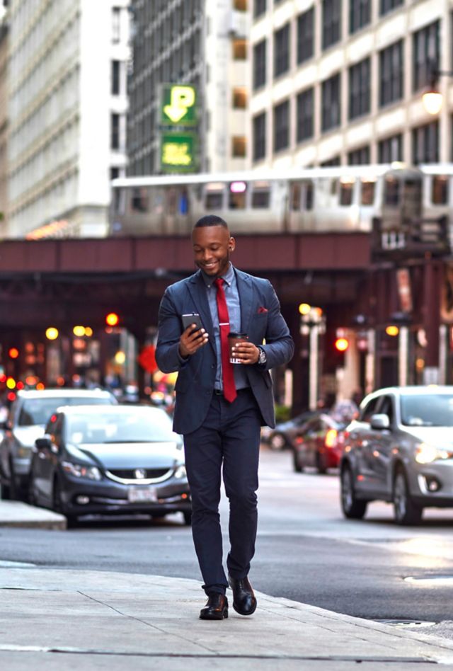 A smiling dark-skinned man walks down a city street looking at a smartphone with coffee in hand. 