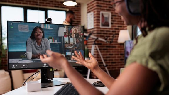 African american woman in casual meeting with startup owner and team in video call from home living room. Job seeker in online interview on personal computer while roommate is relaxing reading a book.