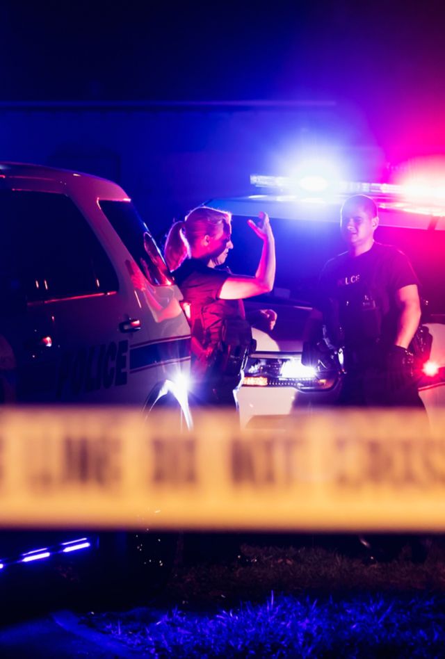 Two multi-ethnic police officers at night wearing bulletproof vests, standing beside police cars with emergency lights flashing, behind cordon tape. The policewoman is in her 40s and her Hispanic, male partner is in his 30s.