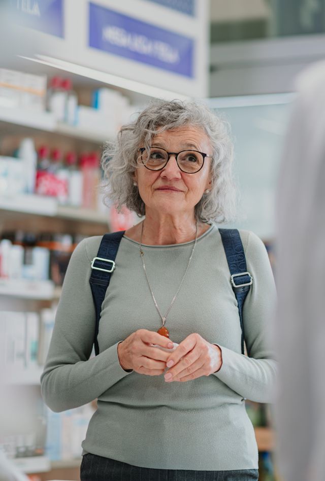 Smiling senior woman with glasses talking to two pharmacists in a brightly lit pharmacy