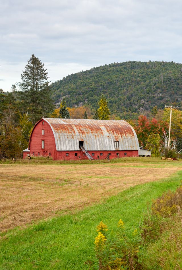 Old red wooden barn with a rusty metal roof along a road in a mountain setting on a cloudy fall day. Ticonderoga, NY, USA.
