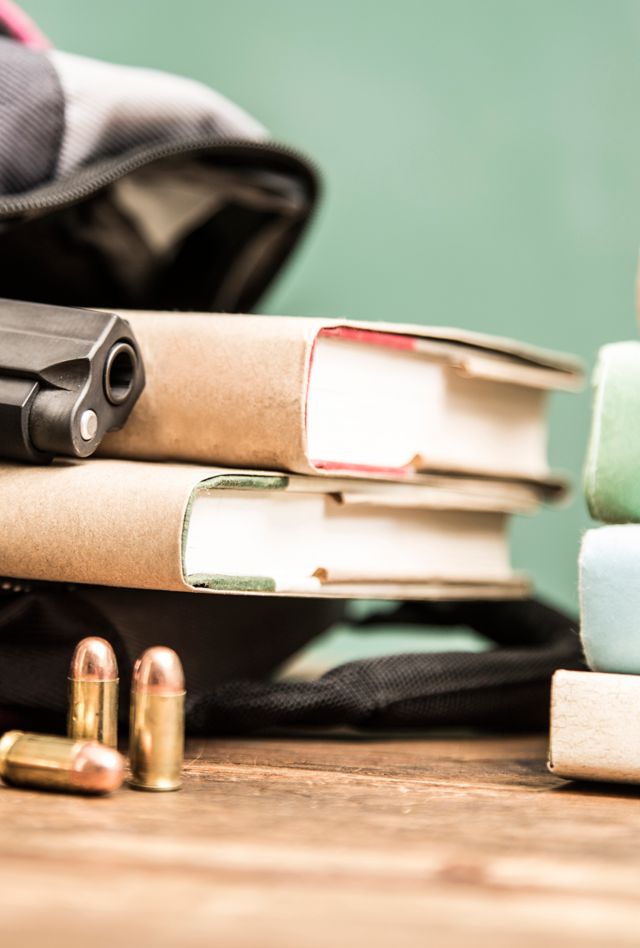 Scene shows gun, bullets and books inside high school student's backpack with book stack and cell phone.  Items lie on school desk with chalkboard background. 