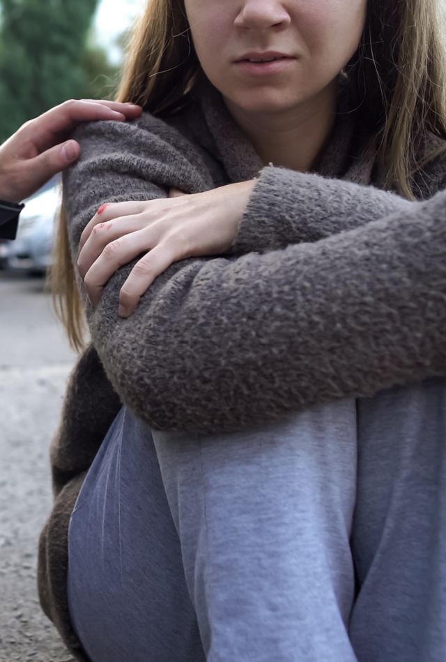 A young woman sitting on the ground with her arms crossed as a person holding a clipboard talks to her