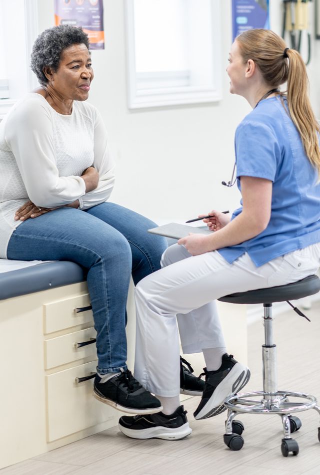 A senior woman of African decent, sits up on an exam table during a medical appointment.  She is dressed casually as she talks with her female doctor who is seated in front of her in scrubs.
