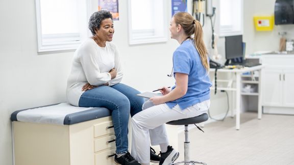 A senior woman of African decent, sits up on an exam table during a medical appointment.  She is dressed casually as she talks with her female doctor who is seated in front of her in scrubs.