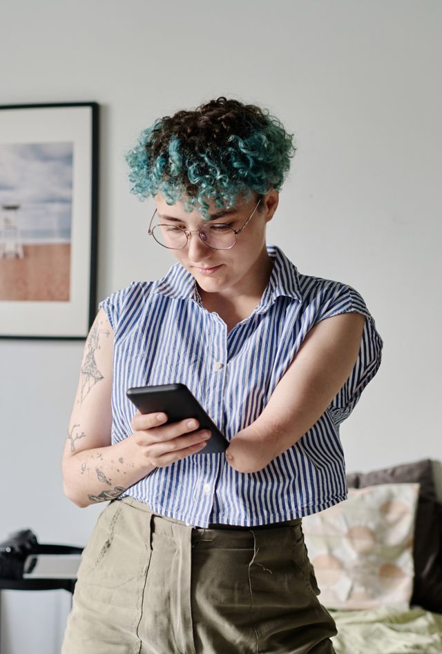 Young woman reading message on her smartphone while standing in the living room. Her hair is dyed teal and her left arm has no forearm.