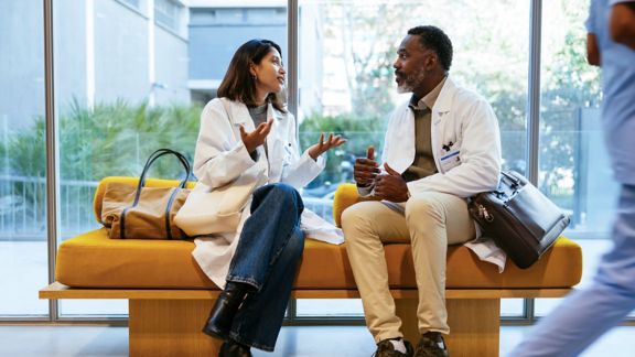 Two medical professionals in conversation, seated on bench in front of windows