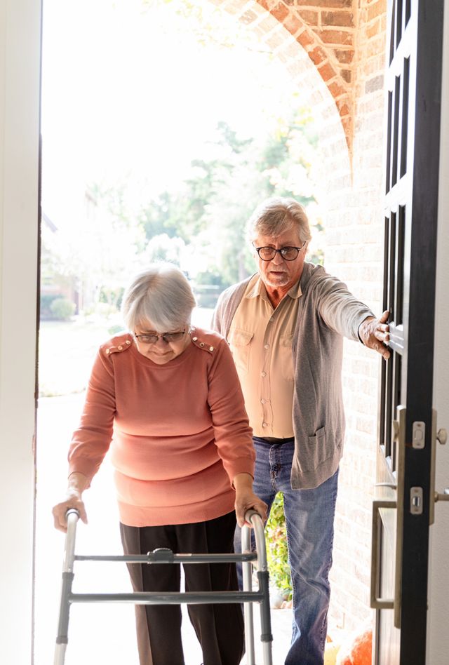 Elderly woman uses a walker, husband helps her inside.