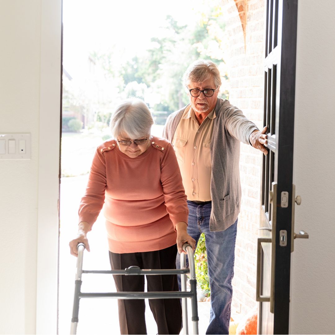 Elderly woman uses a walker, husband helps her inside.