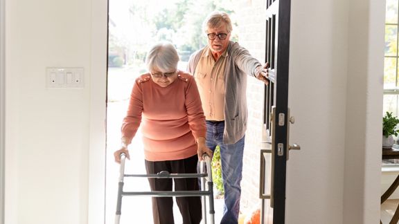 Elderly woman uses a walker, husband helps her inside.
