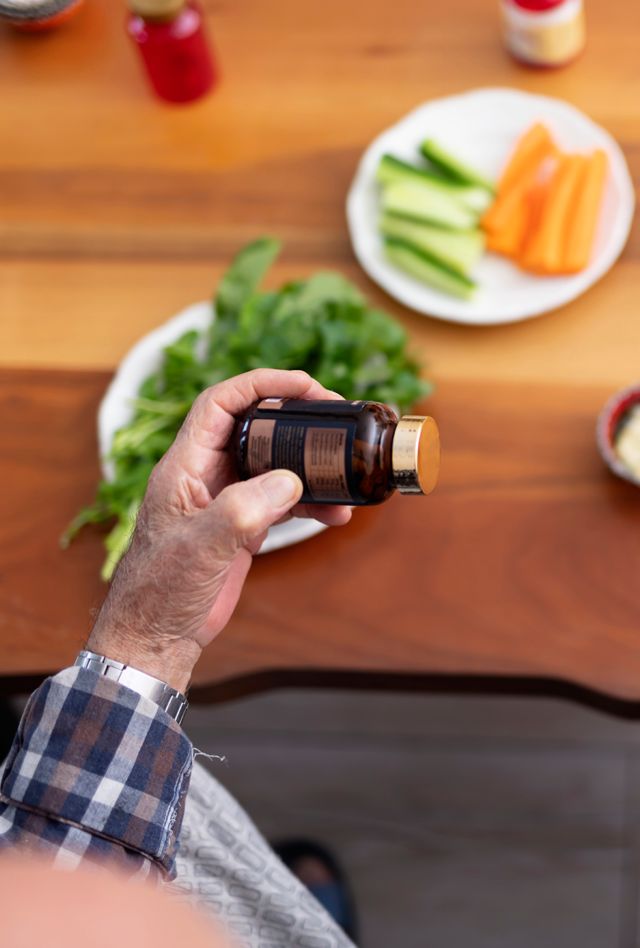 hand holding a pill bottle over a table with vegetables on a plate and other bottles of pills