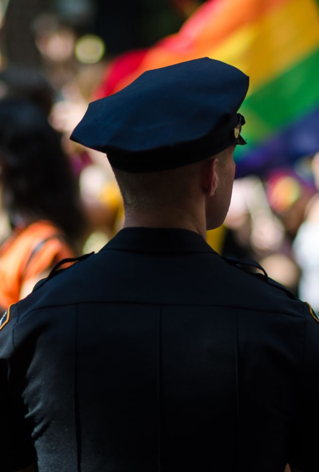 Policeman stands guard in silhouette at pride parade, with rainbow flags in view.
