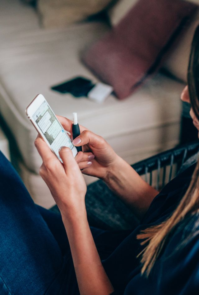 Woman sits in a naturally lit living room while she texts on her smart phone and holds an electronic cigarette.