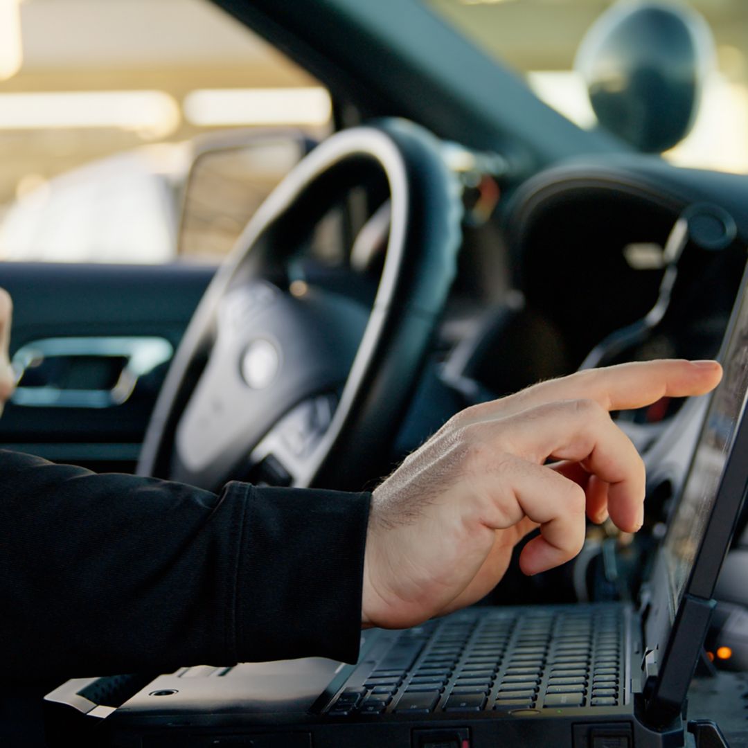 View of the interior of a police car, where a police officer sitting in the driving seat is using a laptop.
