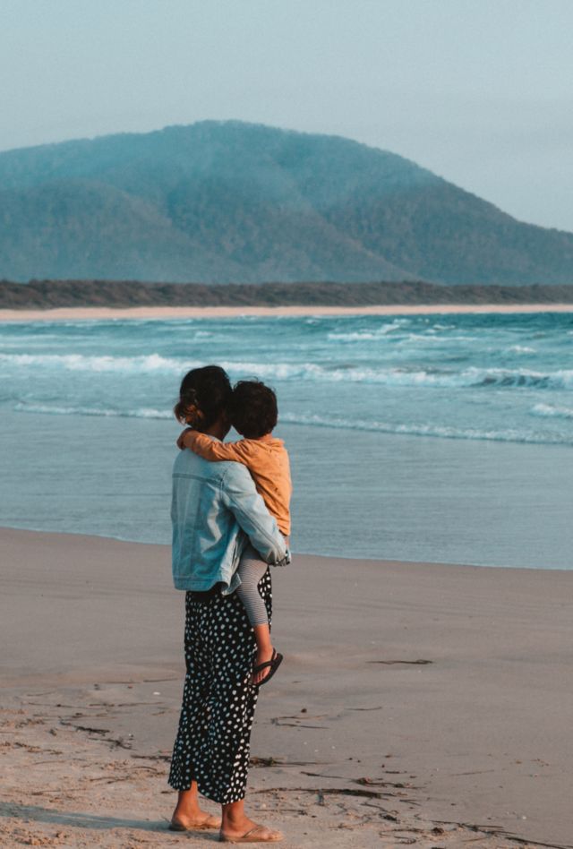 A mother holding a child standing on the sand by the beach