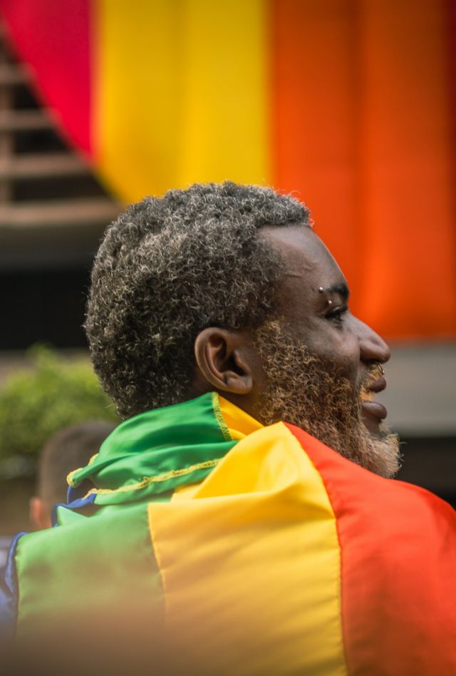 Dark-skinned person with graying hair and beard wrapped in rainbow flag at parade