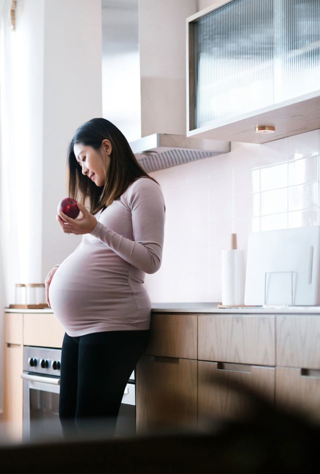 Smiling young Asian pregnant woman touching her belly, eating an apple in kitchen