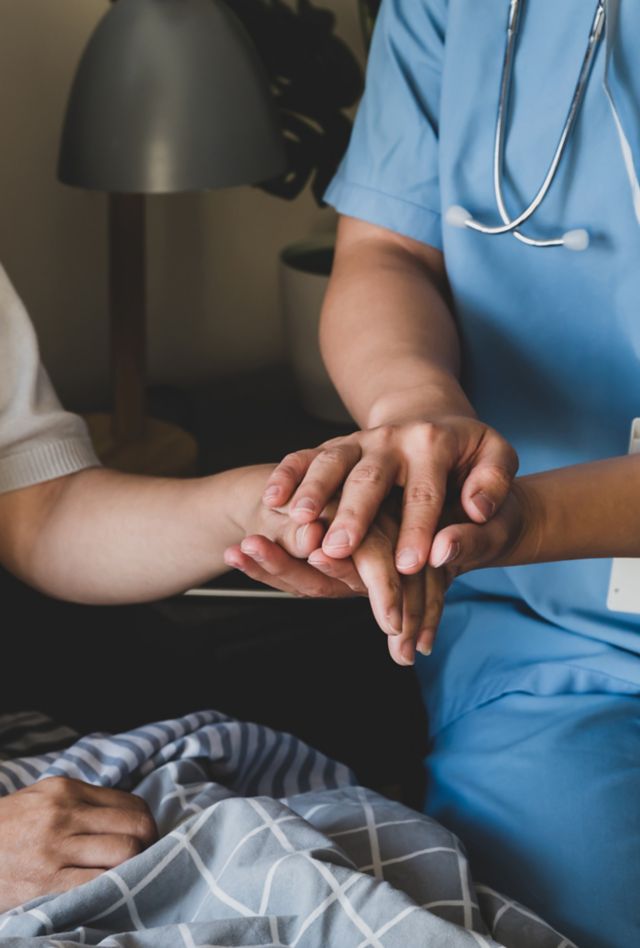 A zoomed shot of a female home caregiver holding the hand of an elderly female patient for moral support in a naturally lit bedroom.