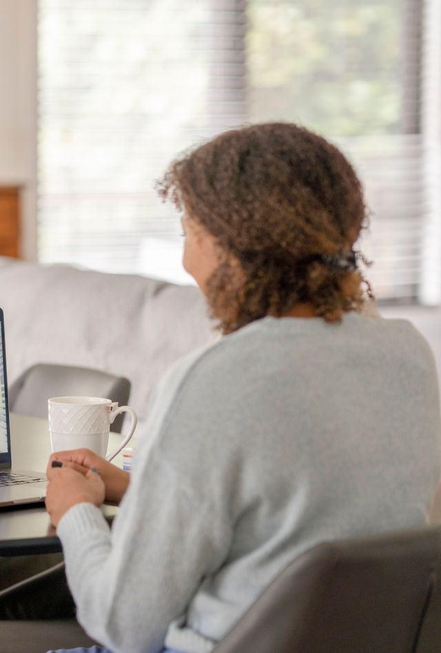 A female patent of African decent meets with her doctor remotely via a video call.  She is sitting at her kitchen table with her laptop out and a cup of coffee on the table with her.  The doctor can be seen on the screen wearing a white lab coat as she talks with her patient.