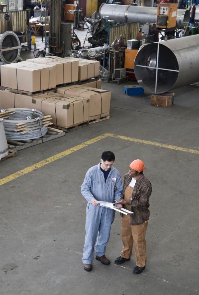 Male Asian American factory worker talking to a female African American factory worker on the floor of a metal manufacturing factory.