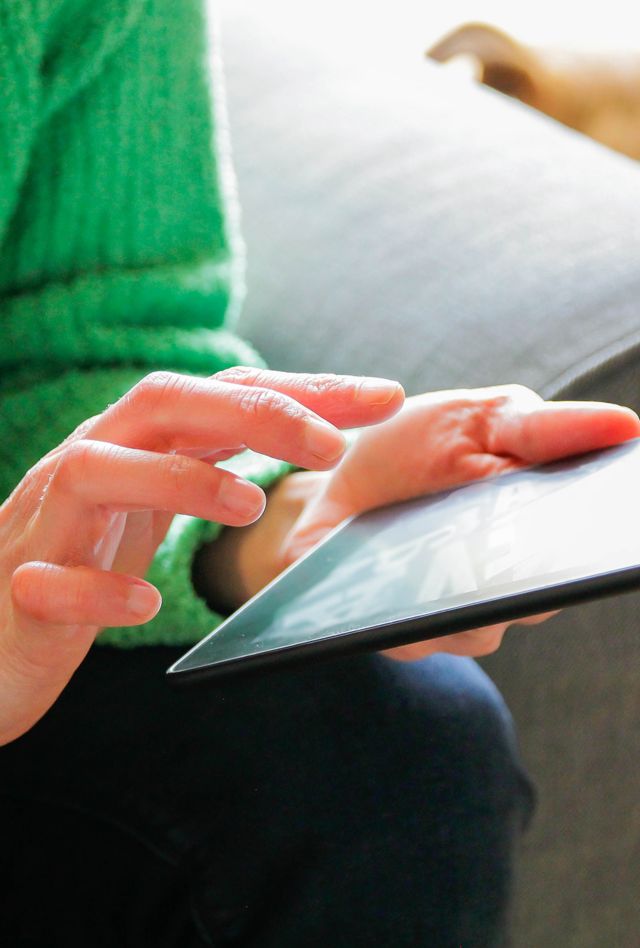 A person in a green sweater uses a tablet while sitting on a couch. A dog is partially visible in the background.