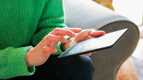 A person in a green sweater uses a tablet while sitting on a couch. A dog is partially visible in the background.