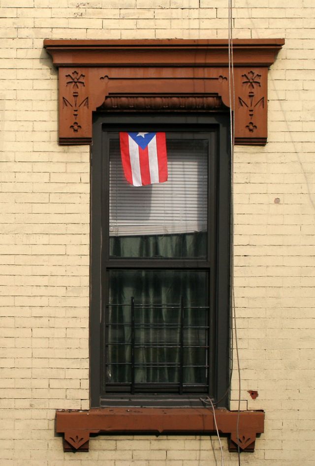 street view of brick wall with two windows. One has a air conditioning unit, the other has blinds, a curtain, and a Puerto Rican flag.