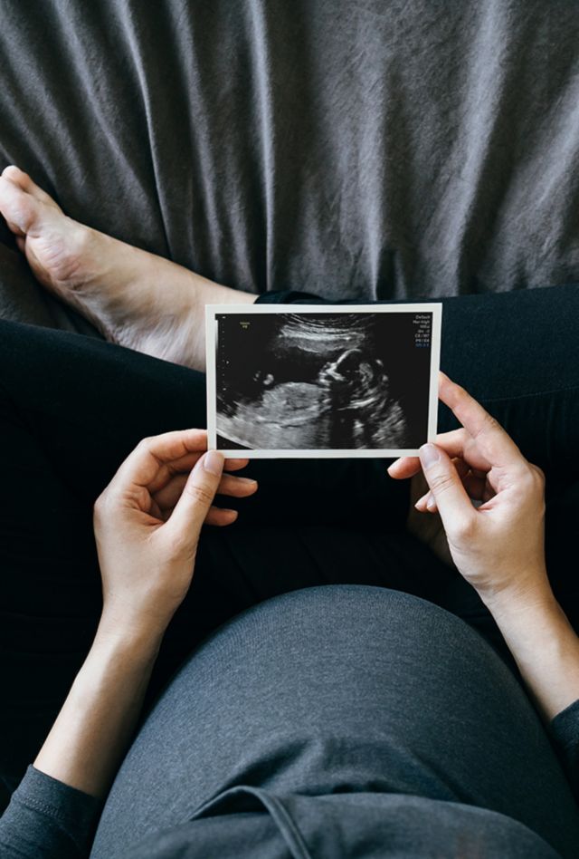 High angle shot of pregnant woman holding an ultrasound scan photo in front of her baby bump, sitting on bed at home.