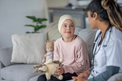 A young girl wearing a pink sweater and headscarf sits on a sofa, holding a plush toy while a smiling female doctor talks with her. A warm, supportive medical consultation at home.
