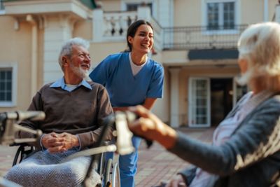 A cheerful caregiver is outdoors in the yard of the nursing home with elderly patients having a laugh with them.