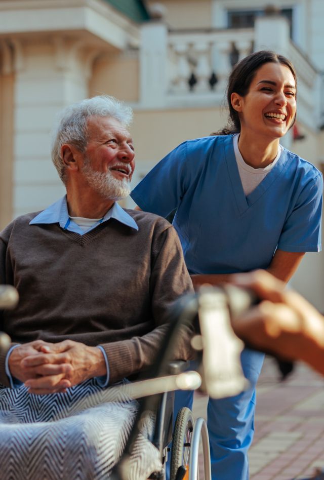 A cheerful caregiver is outdoors in the yard of the nursing home with elderly patients having a laugh with them.