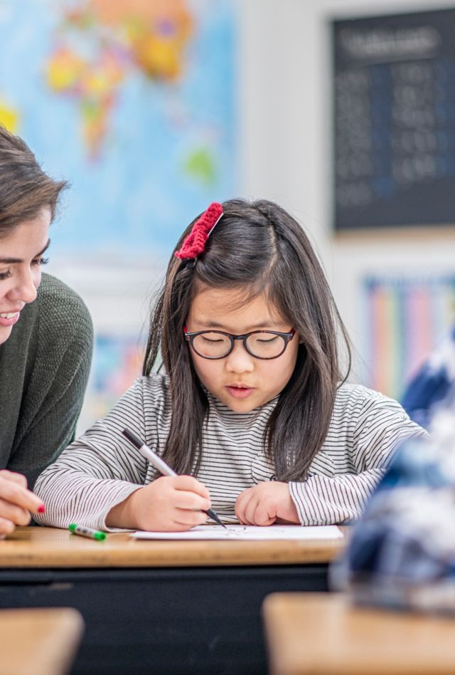 A teacher bends down beside the desk of a female Asian student as she helps her with her work.  Her multi-ethnic peers can be seen working away quietly and individually at their own desks.  They are all dressed casually and have neutral expressions on their faces as they focus on their tasks.