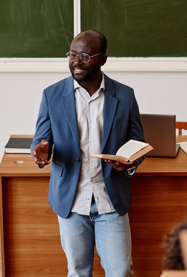 Dark-skinned teacher standing with book and lecturing to students in university classroom setting