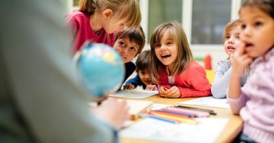 Preschool teacher teaching her children about geography. Using globe and asking the questions. Children sitting by the table and listen teacher carefully. Models in this shot are part of real kindergarten group and their teacher.