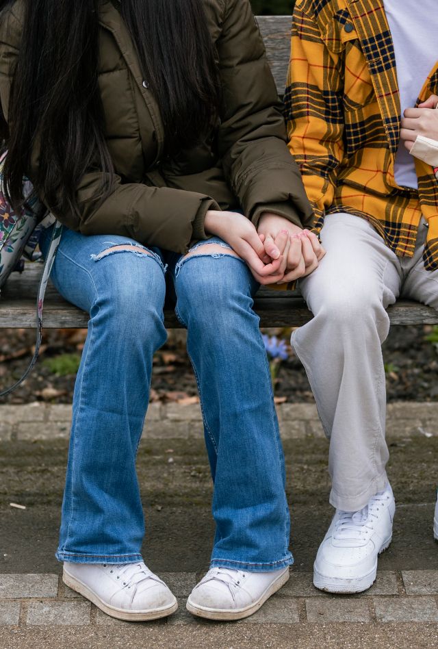 A close-up shot of an unrecognizable teenage couple sitting on a wooden bench in a public park in Wallsend, North East England. The main focus is the midsection of their bodies and the couple holding hands.