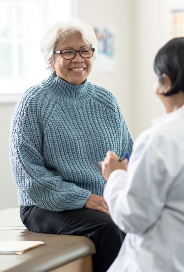 A senior woman sits up on the exam table at a doctors appointment.  She is dressed casually in a blue sweater and has a smile on her face as she glances at the doctor.  Her female doctor is wearing a white lab coat and seated in front of her as she takes notes on a tablet.